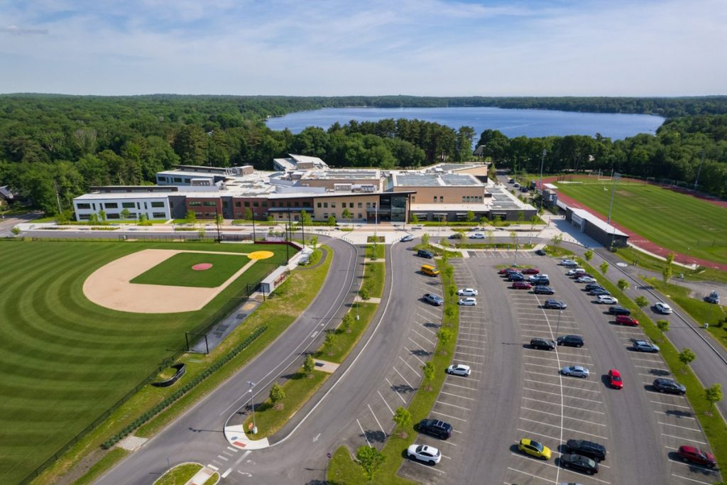 Aerial view of the Sharon High School property prominently showing the building, parking lot and baseball field.  Behind it, Lake Massapoag in the distance.